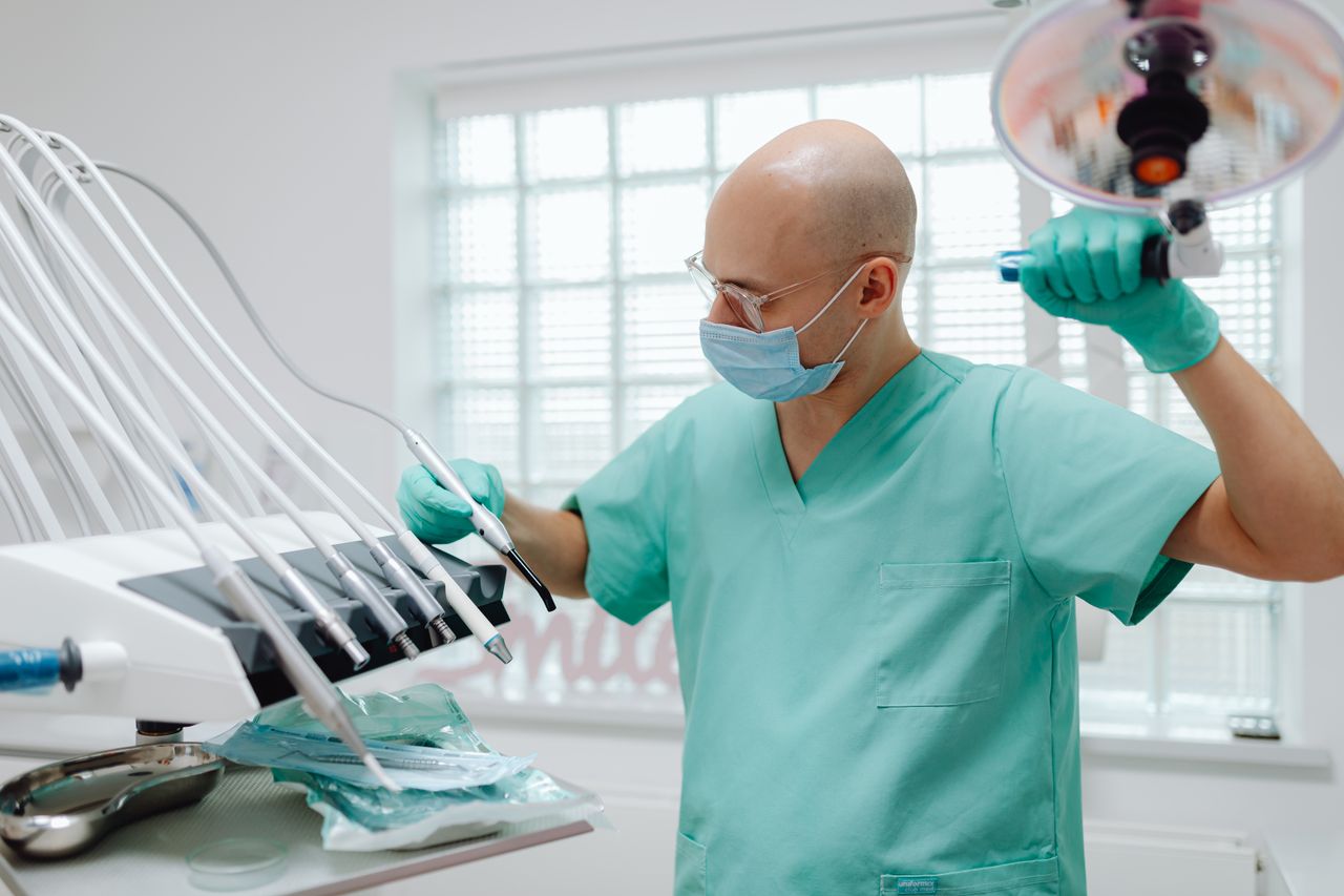 A bald dentist wearing green scrubs and a face mask adjusting dental equipment in a clinic setting.